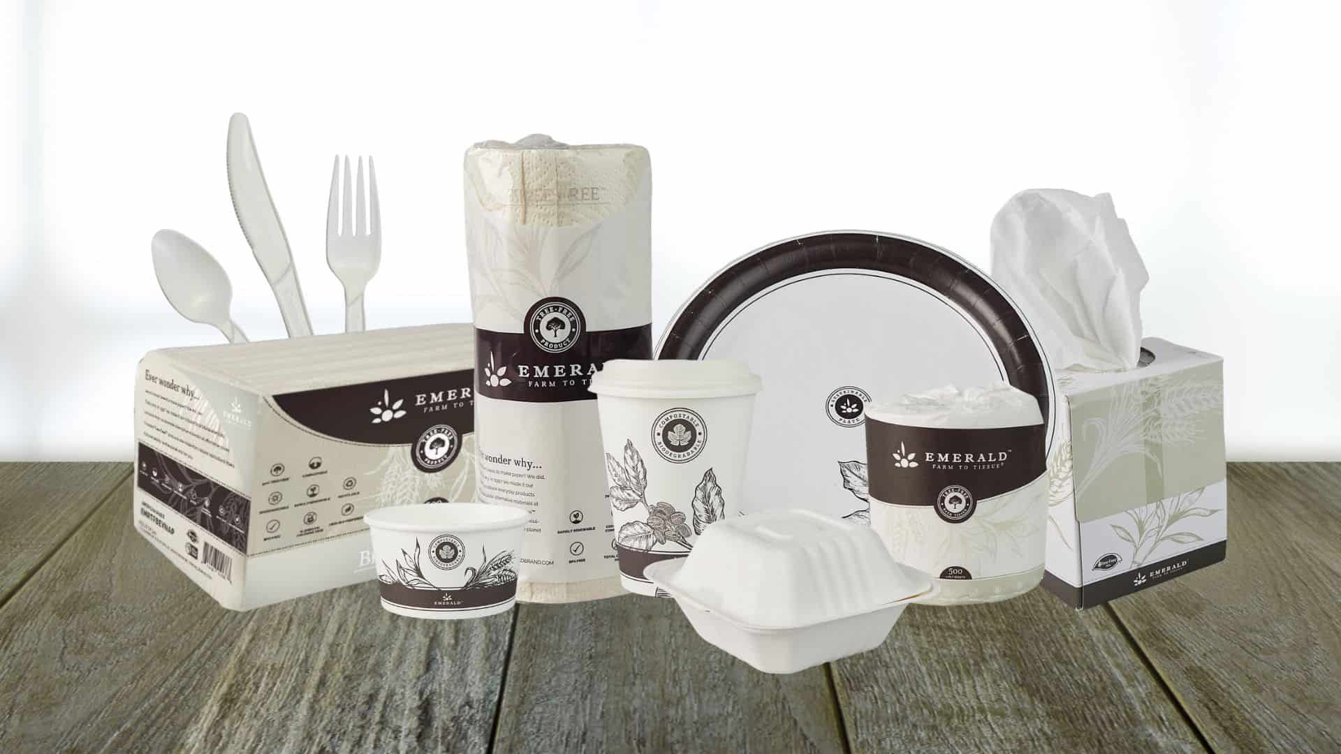 A collection of eco-friendly disposable tableware and tissues includes plates, cups, cutlery, a napkin pack, and two tissue boxes. All items feature brown and white packaging with a green theme, displayed on a wooden surface.